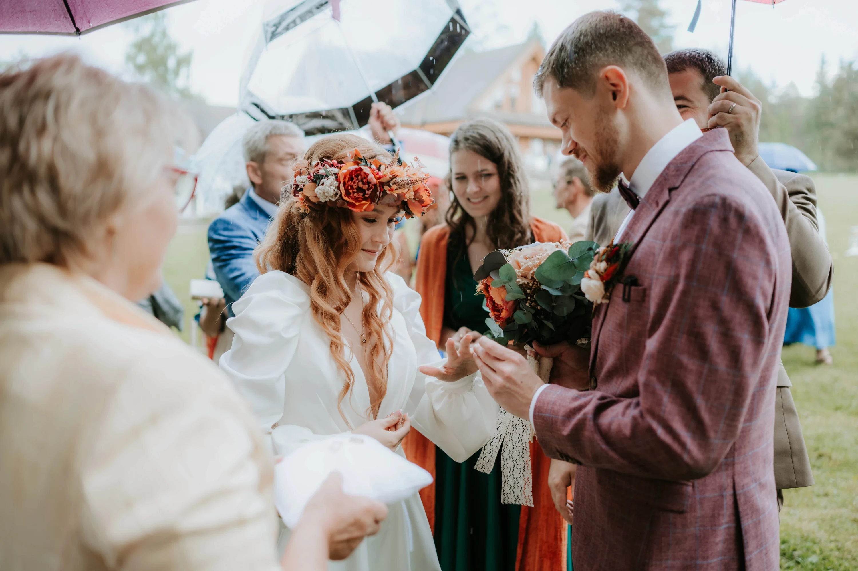 Nevesta navlieka ženíchovi obrúčku počas obradu v daždi, vedľa stojí žena s vankúšikom.