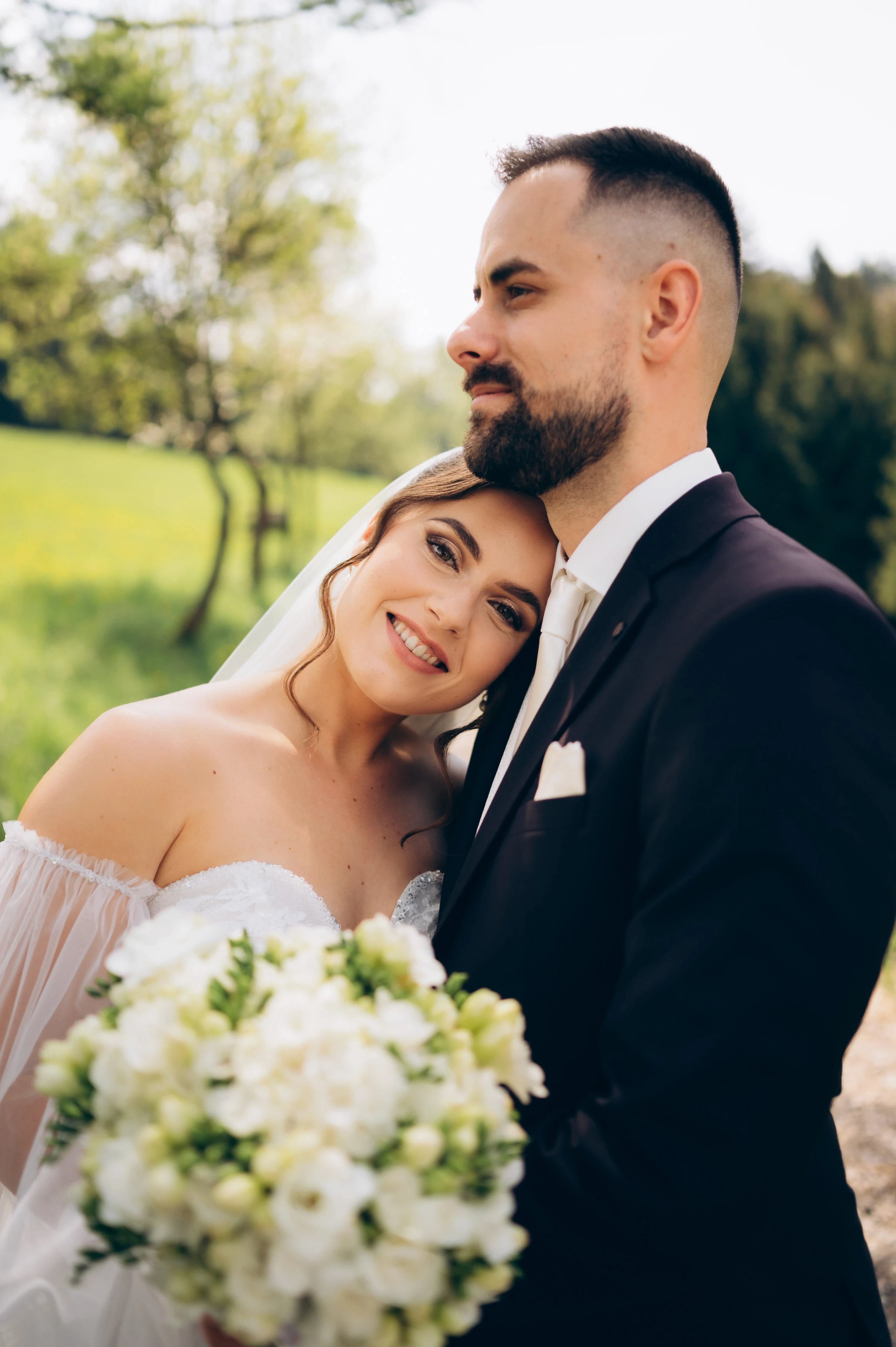 Nevesta s bielou kyticou sa opiera o plece ženícha na okraji zelenej lúky.