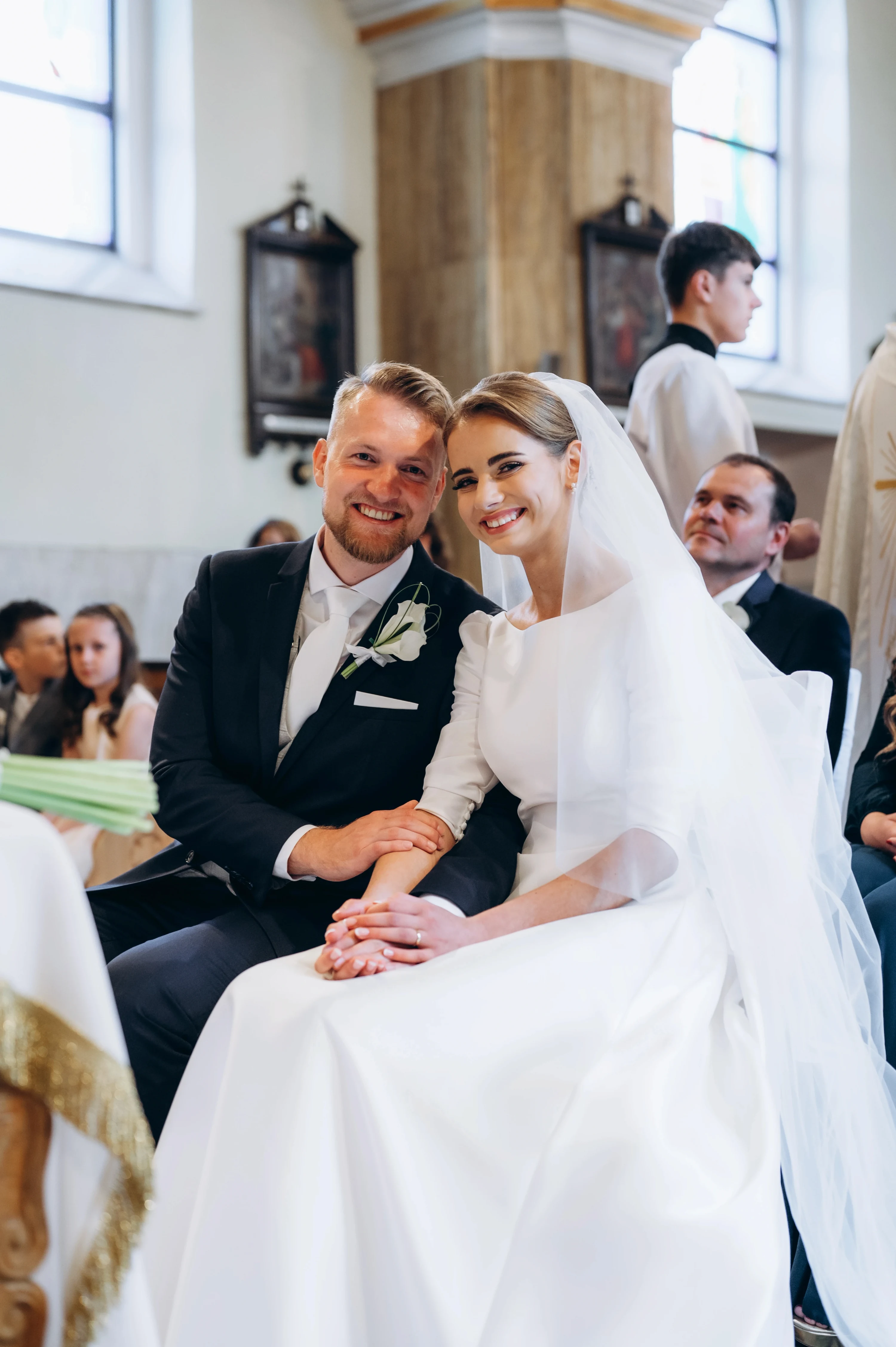 Nevesta a ženích sedia počas obradu v lavici a spolu sa usmievajú do fotoaparátu.
