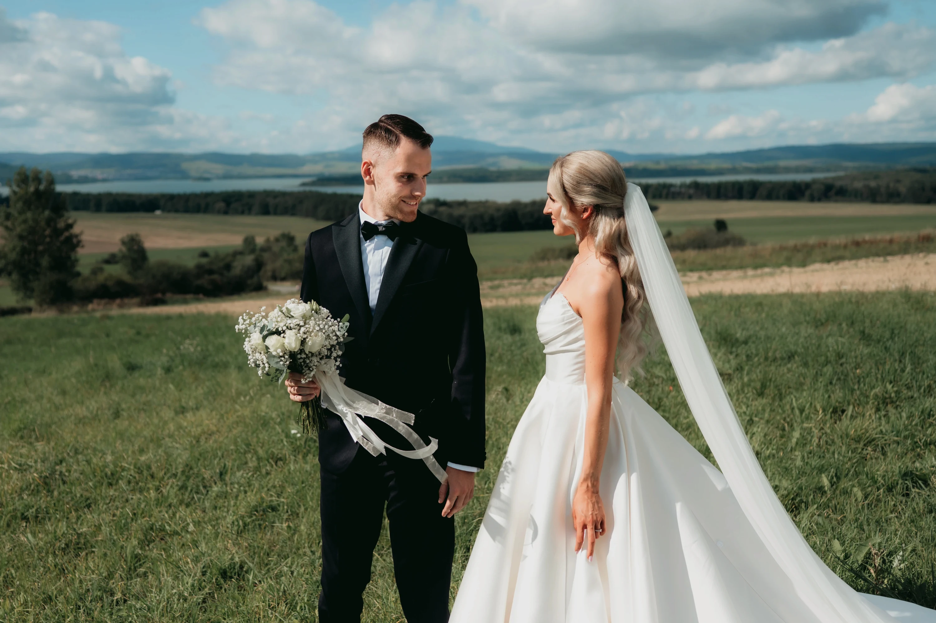Nevesta so závojom a ženích s kyticou stoja proti sebe na lúke nad širokým údolím.