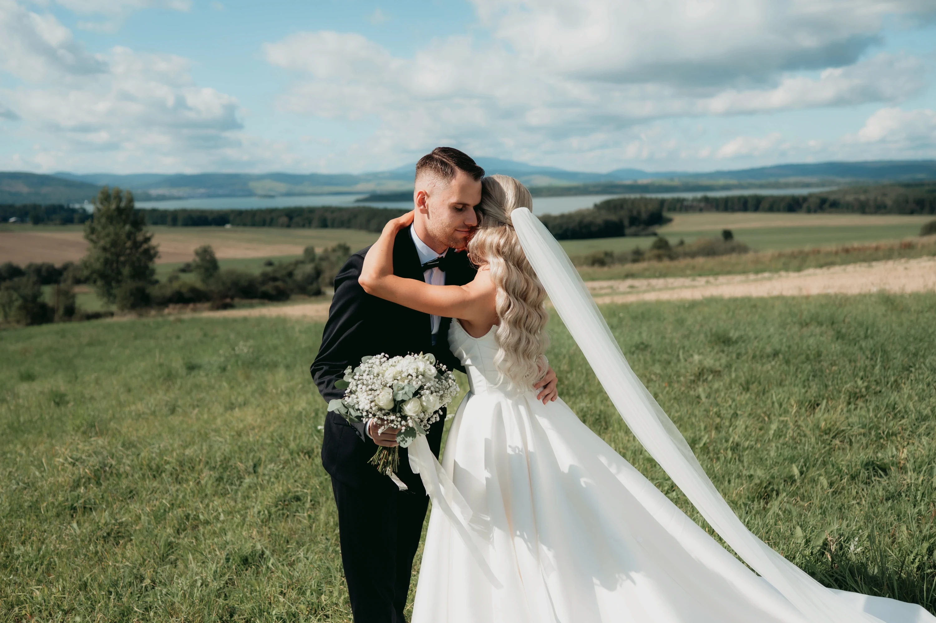 Ženích bozkáva nevestu s dlhým závojom a kyticou na lúke.