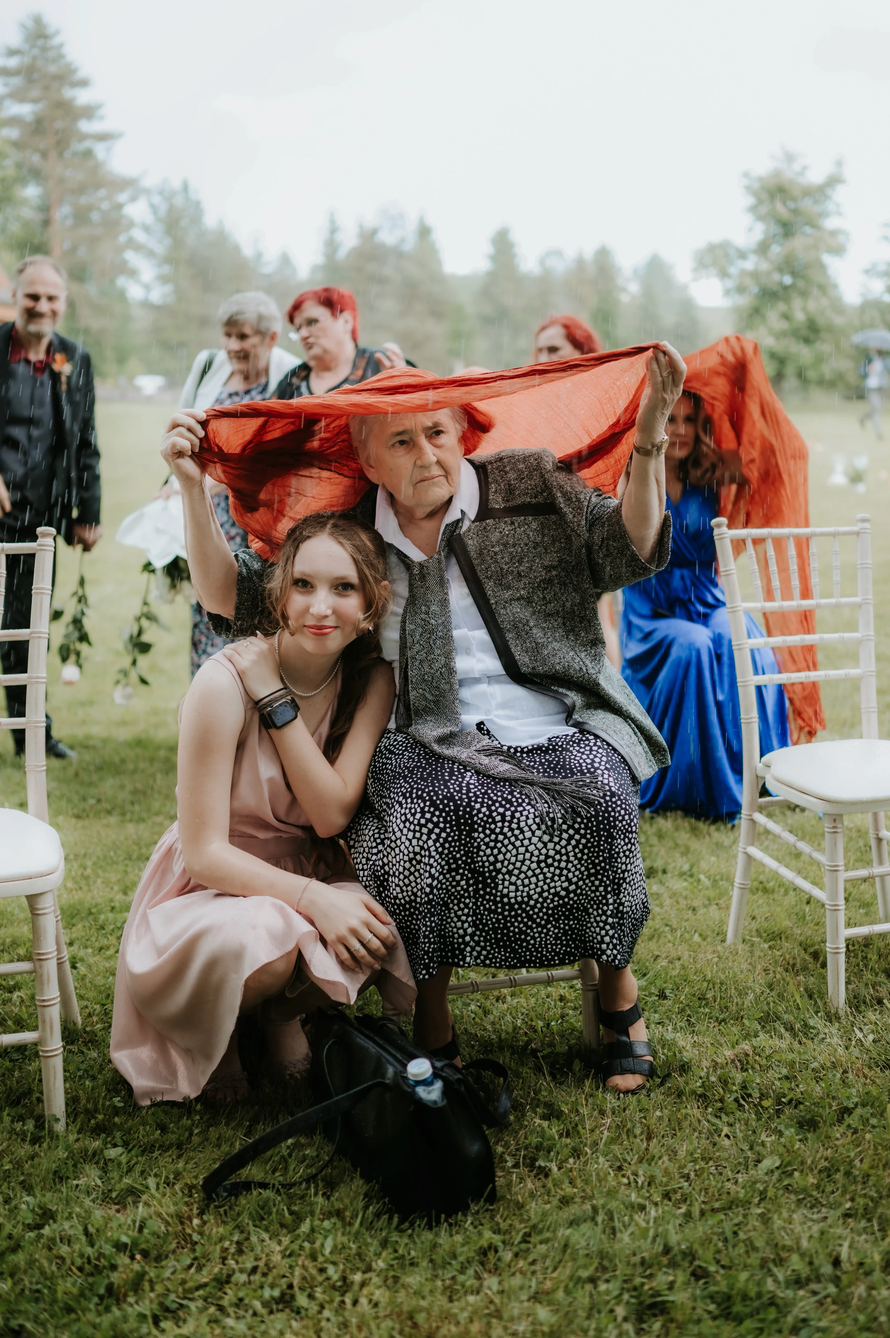 Mladá žena kľačí pri staršom pánovi, nad ktorým hostia držia oranžovú látku proti dažďu.