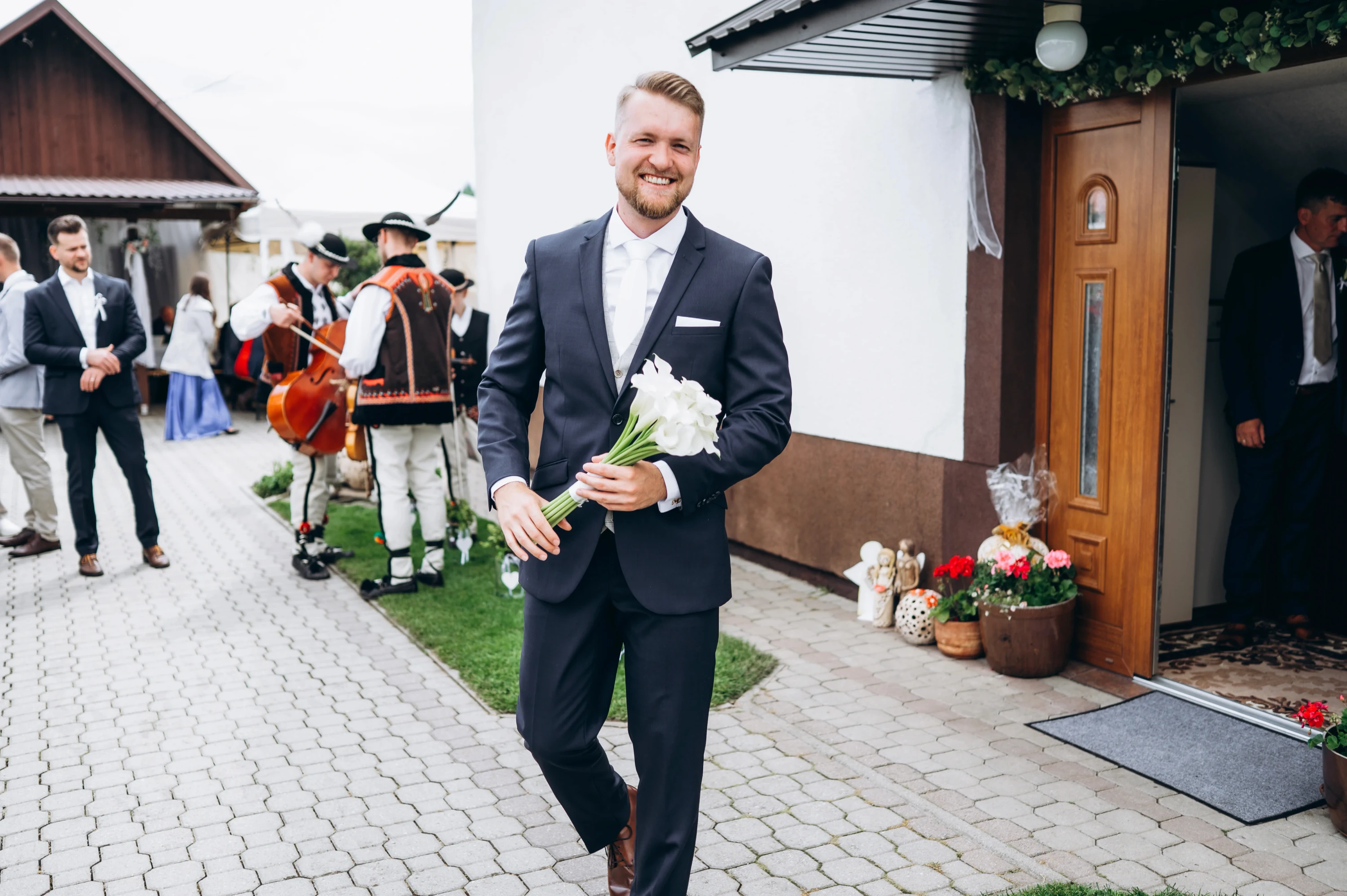 Ženích s kyticou bielych kvetov kráča po dvore, v pozadí hrá ľudová hudba v krojoch.