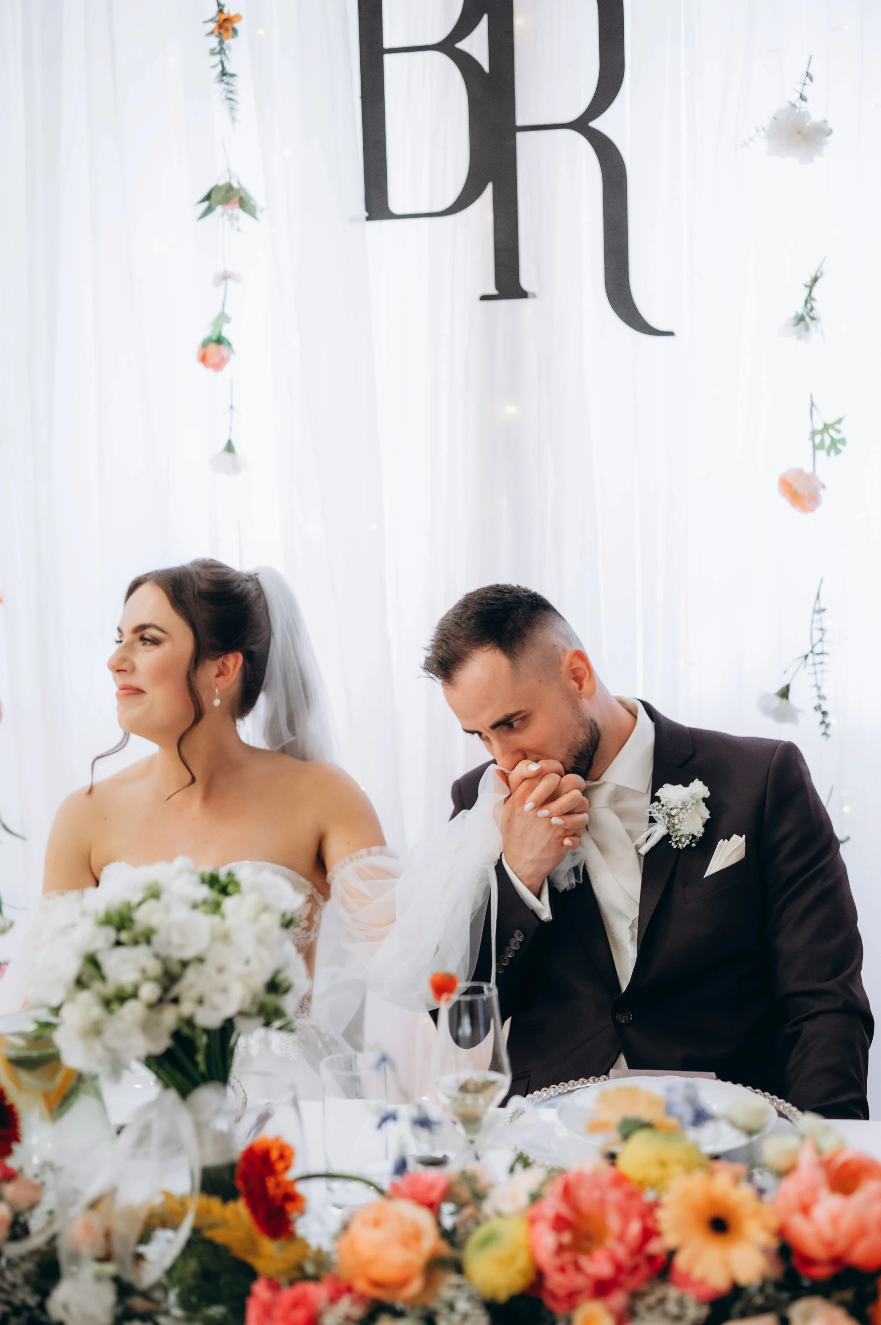 Nevesta a ženích sedia pri stole medzi kvetinovou výzdobou.
