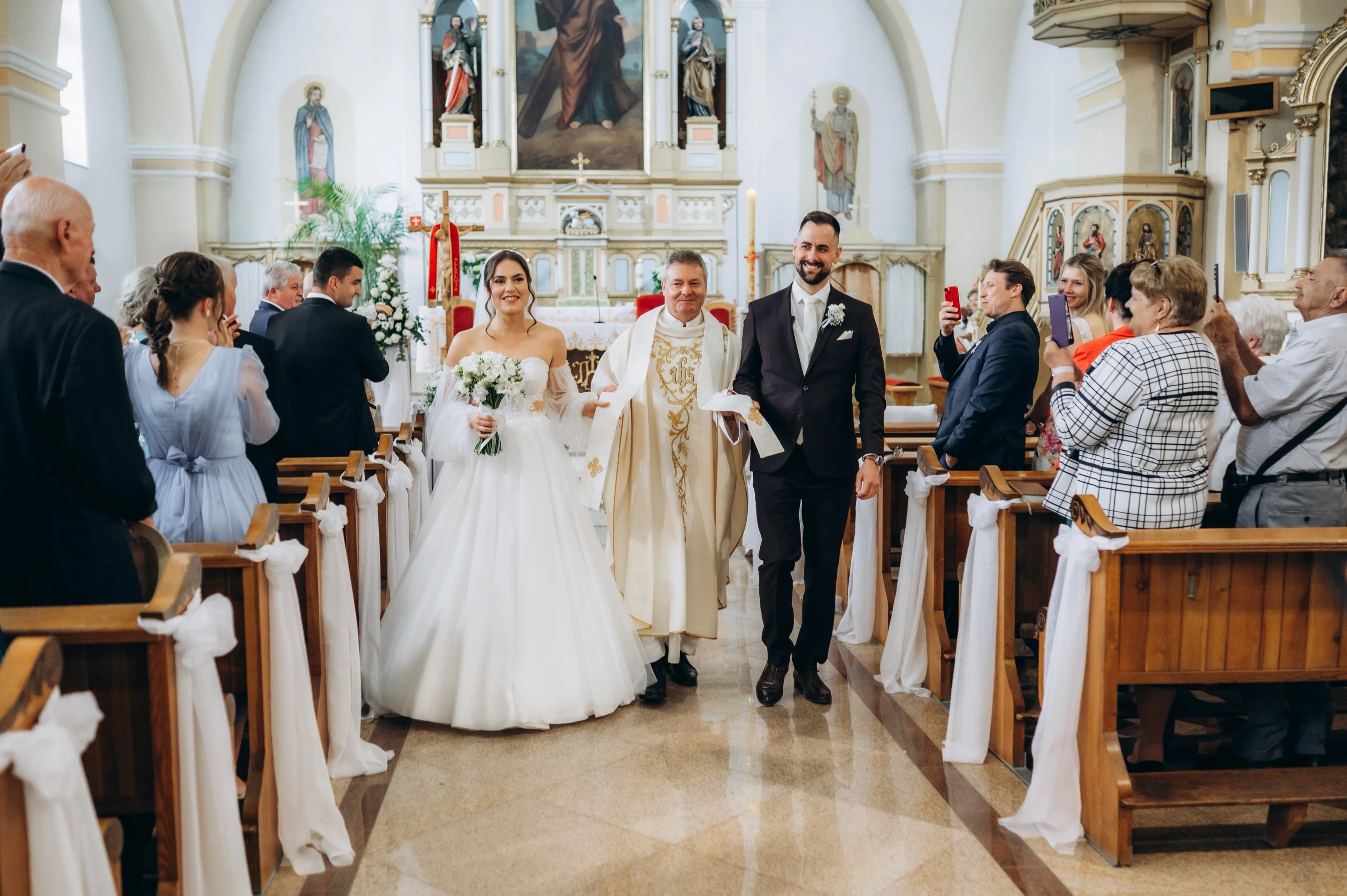 Novomanželia kráčajú kostolnou uličkou od oltára, kňaz ide za nimi a hostia tlieskajú.