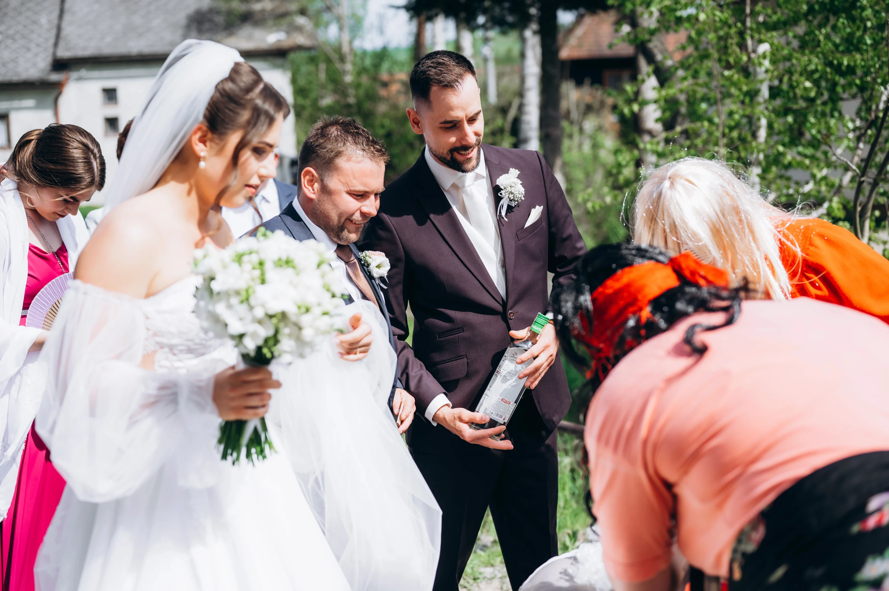Nevesta s kyticou a ženích s fľašou v ruke sledujú zohnutú ženu počas svadobného zvyku.