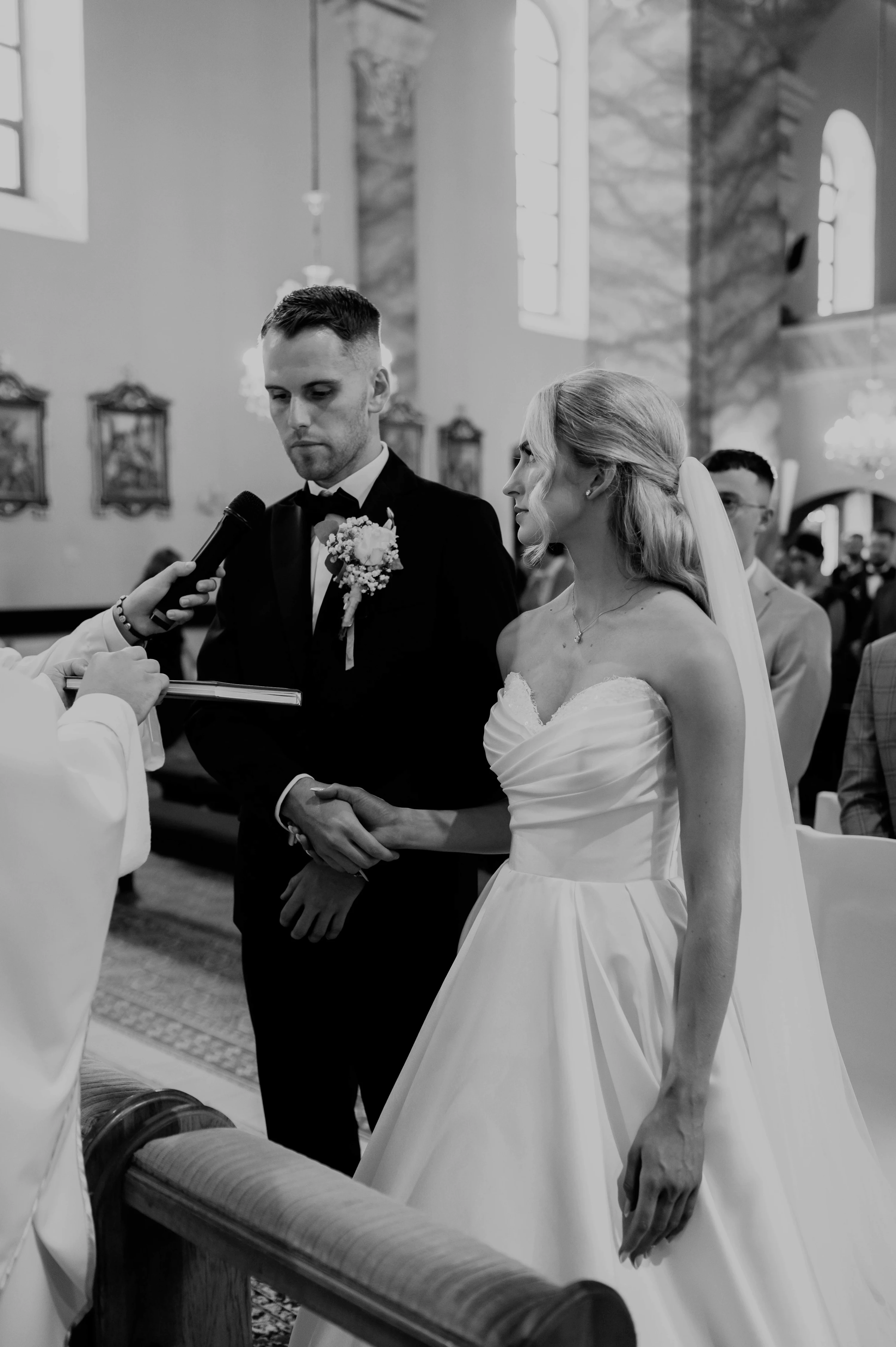 Nevesta a ženích si počas obradu držia ruky, kňaz k nim nakláňa mikrofón.