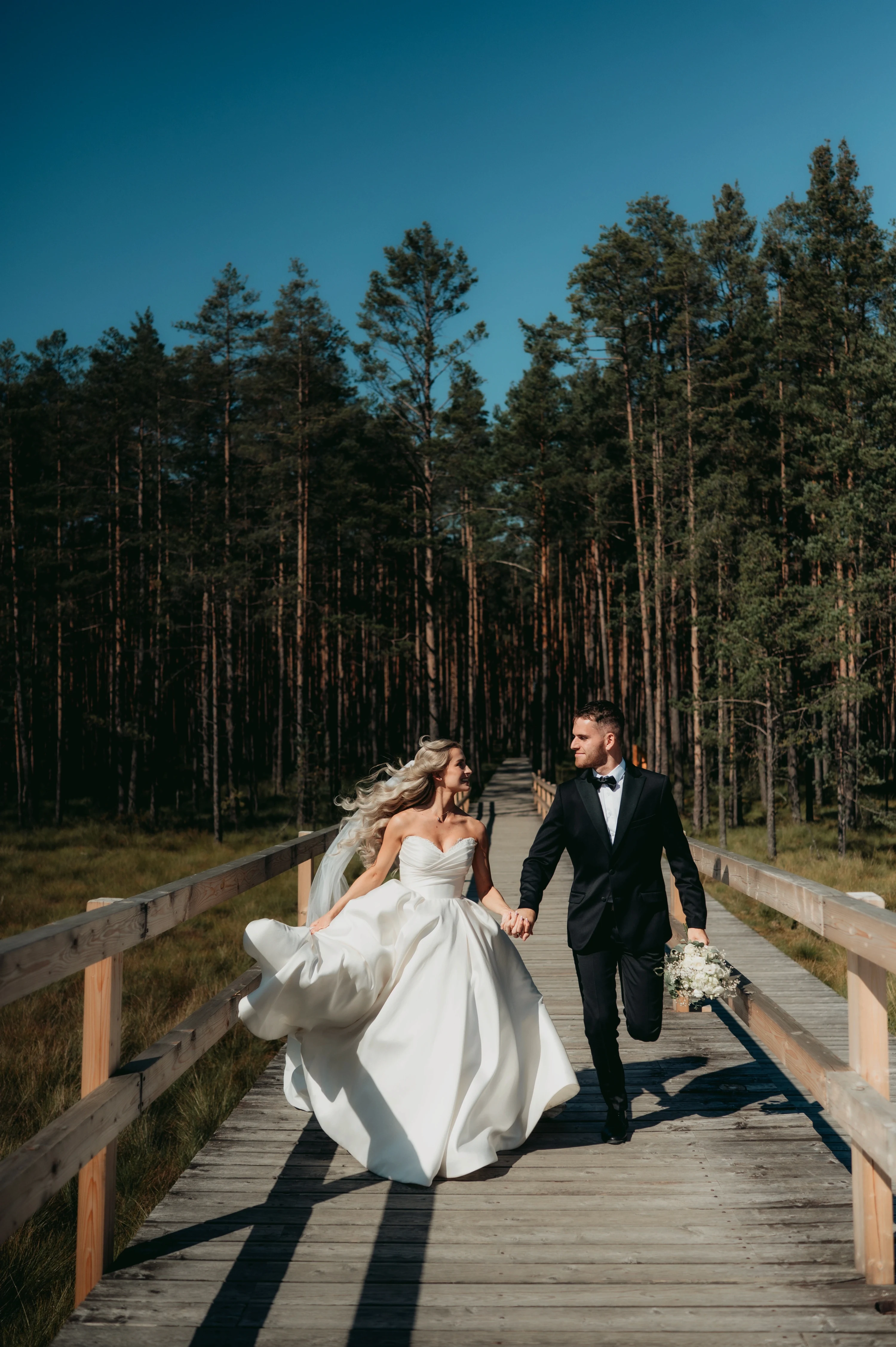 Novomanželia kráčajú po drevenej lávke cez borovicový les pod sýtomodrou oblohou.