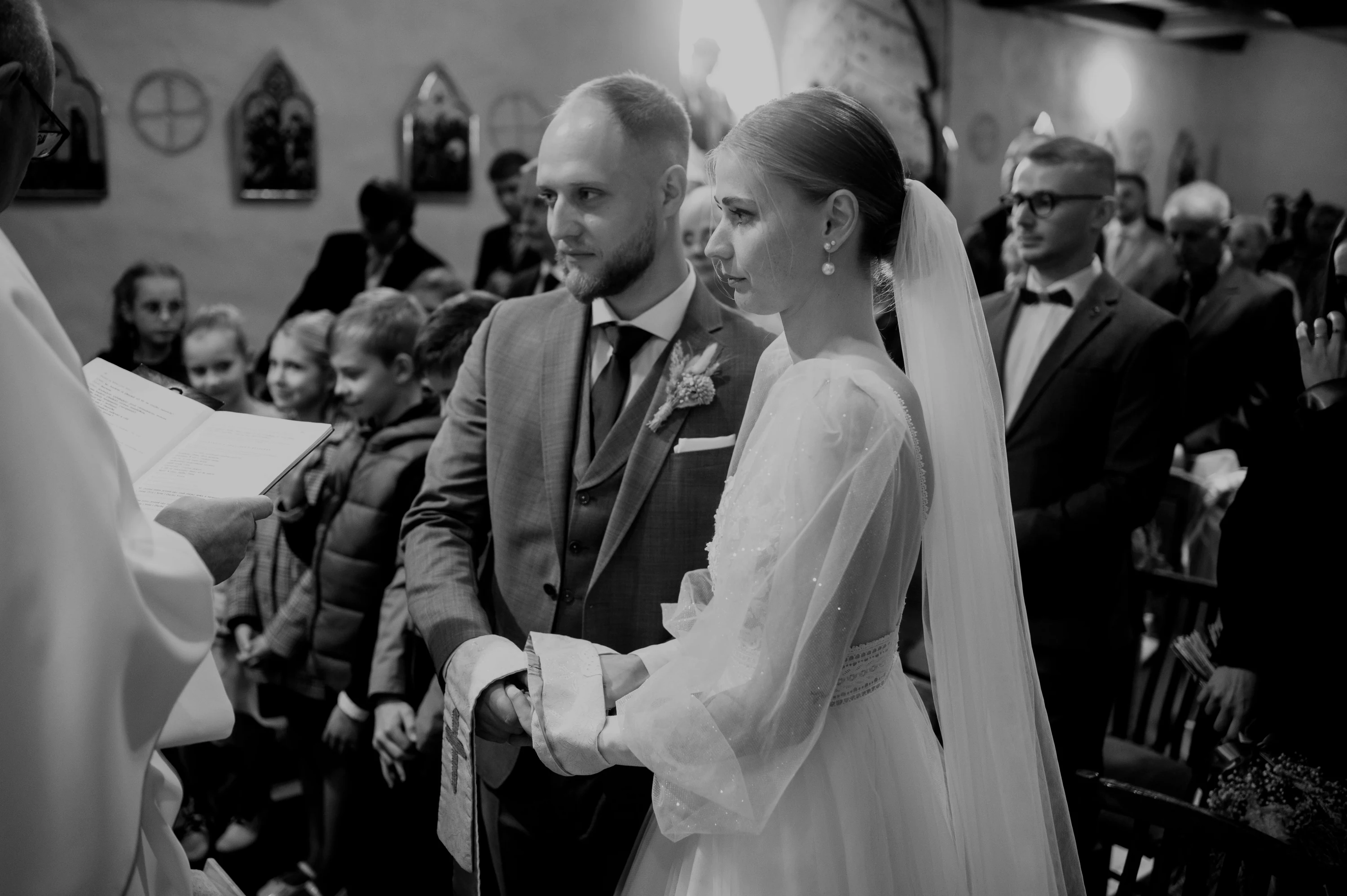 Nevesta so ženíchom stoja pred kňazom, v laviciach za nimi prizerajú deti.