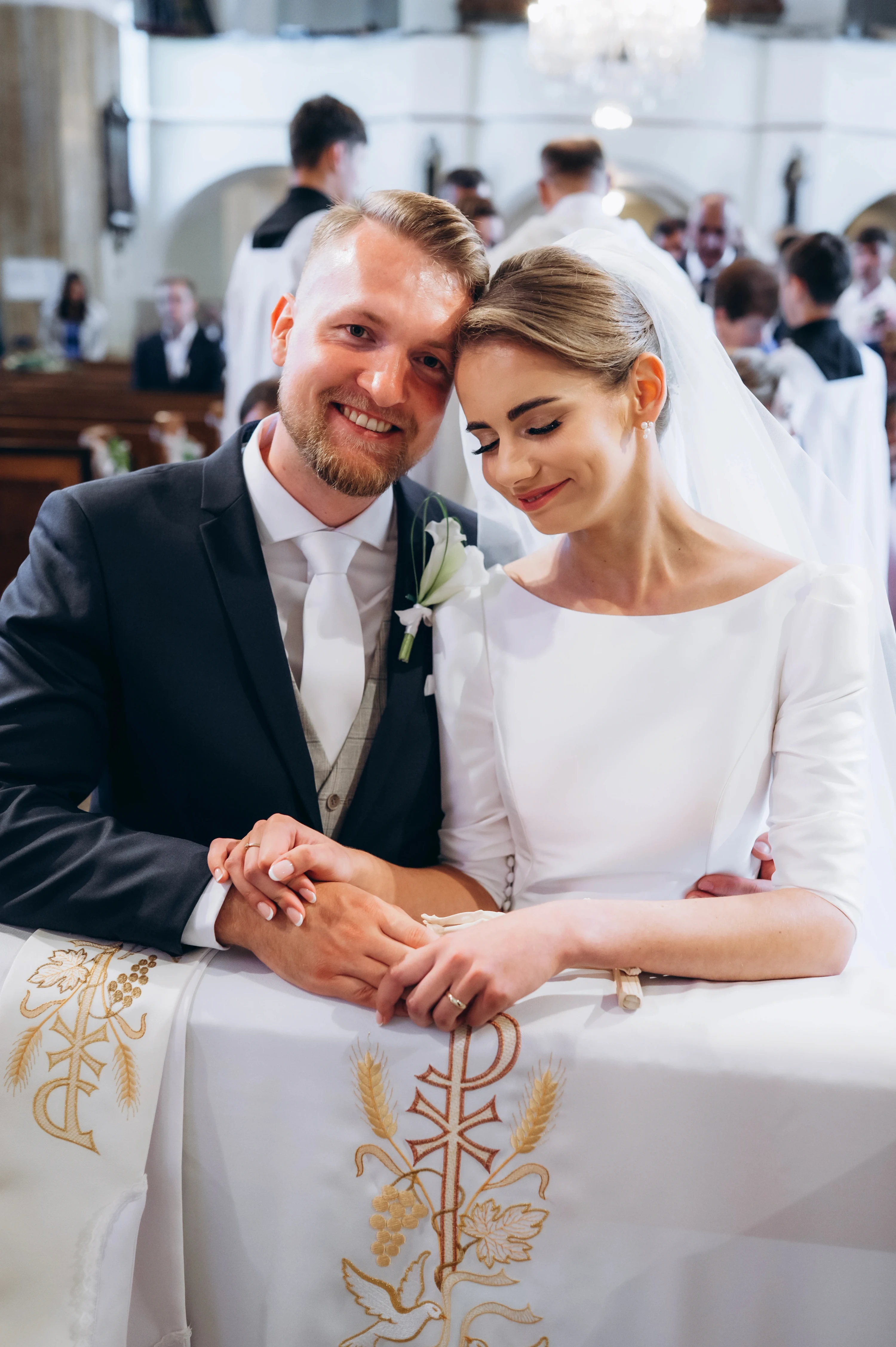 Novomanželia s prepletenými rukami kľačia pri vyšívanej oltárnej plachte a usmievajú sa.