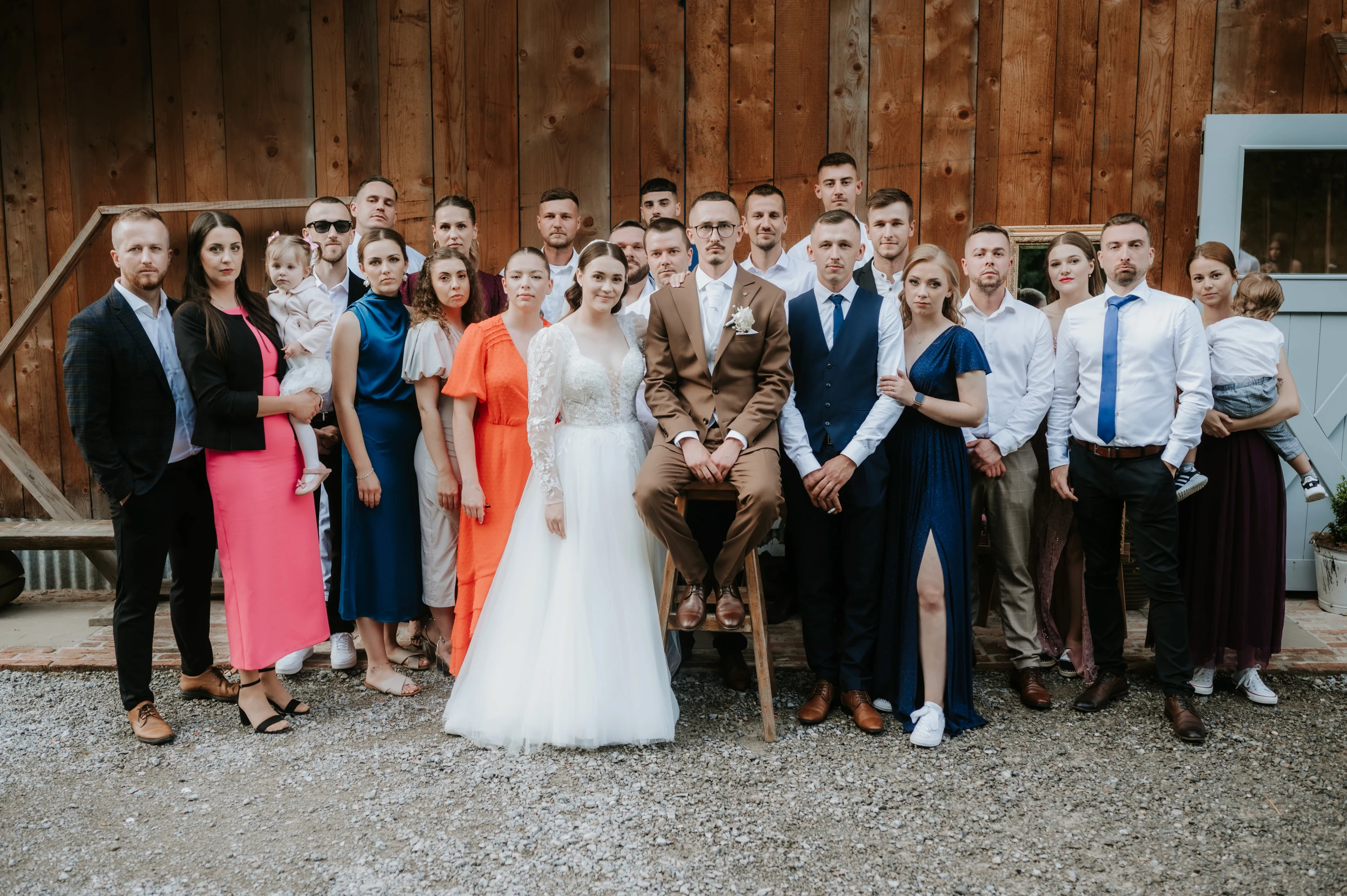 Skupinová fotografia manželov so všetkými hosťami pred drevenou stavbou.